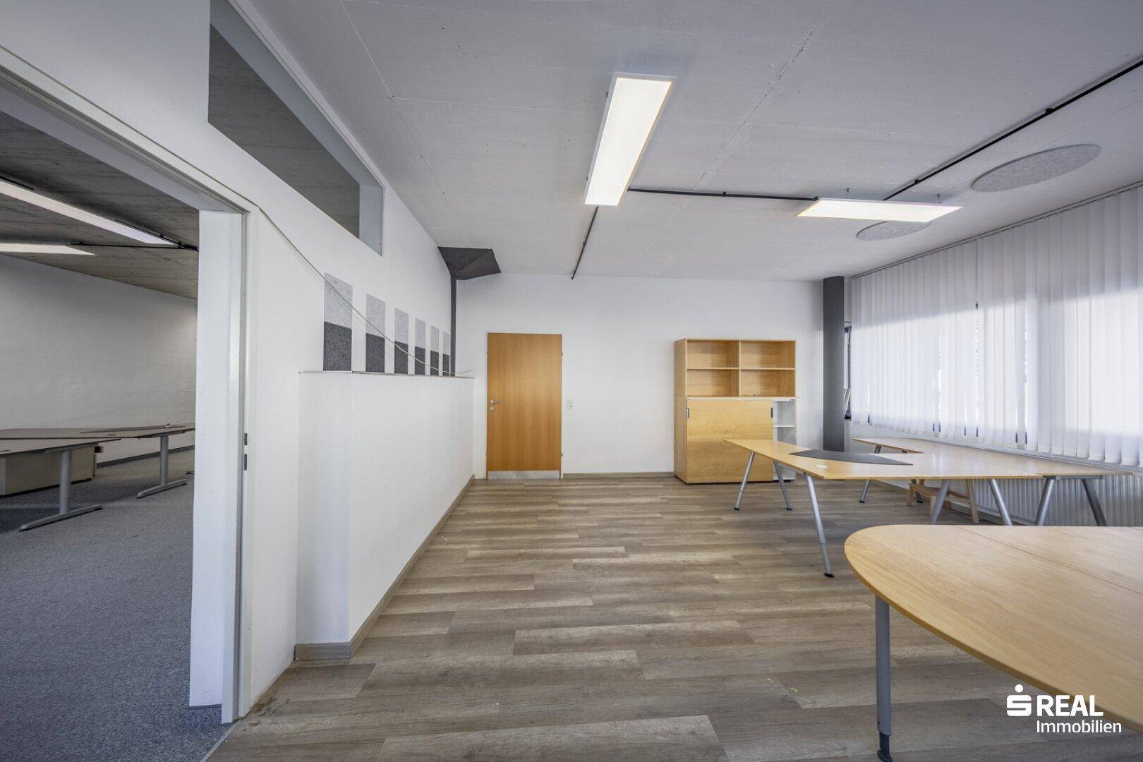 Heller Büroraum mit Holzoptik-Laminatboden, großen Fenstern und Zugang zu einem weiteren Bereich.