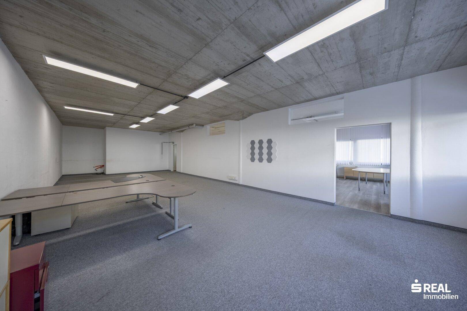Geräumiger Bürobereich mit Teppichboden, Sichtbetondecke und Klimaanlage, offen zu einem Nebenraum.