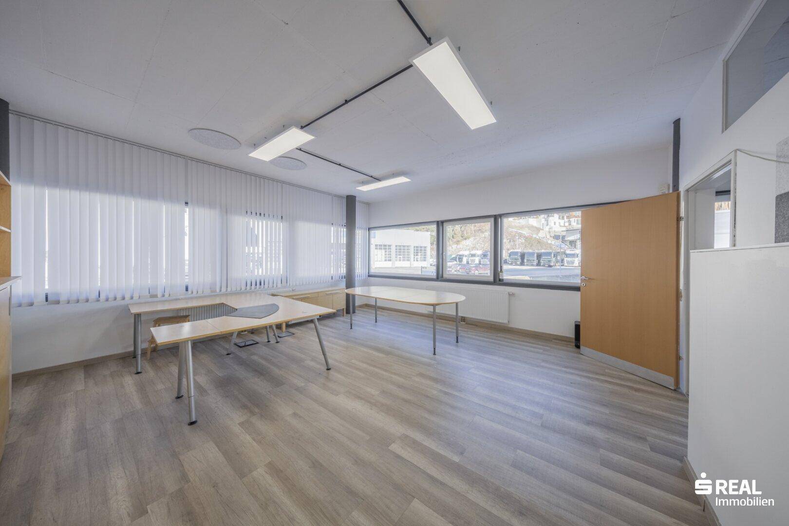 Heller Büroraum mit großen Fenstern, Holzoptik-Laminatboden und Blick auf die Umgebung.