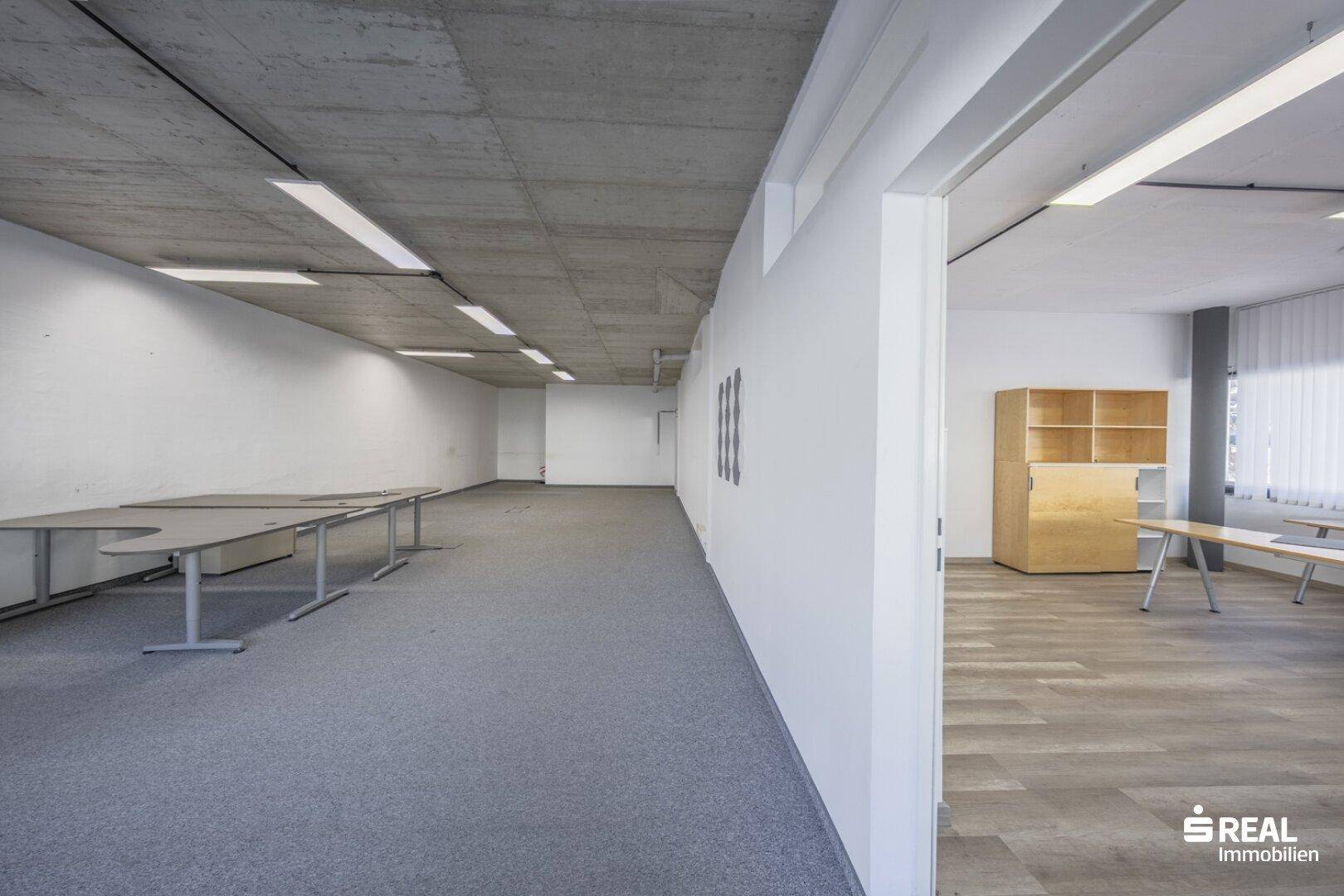 Langer Bürogang mit Teppichboden und Blick in einen angrenzenden Raum mit Holzoptik-Boden.
