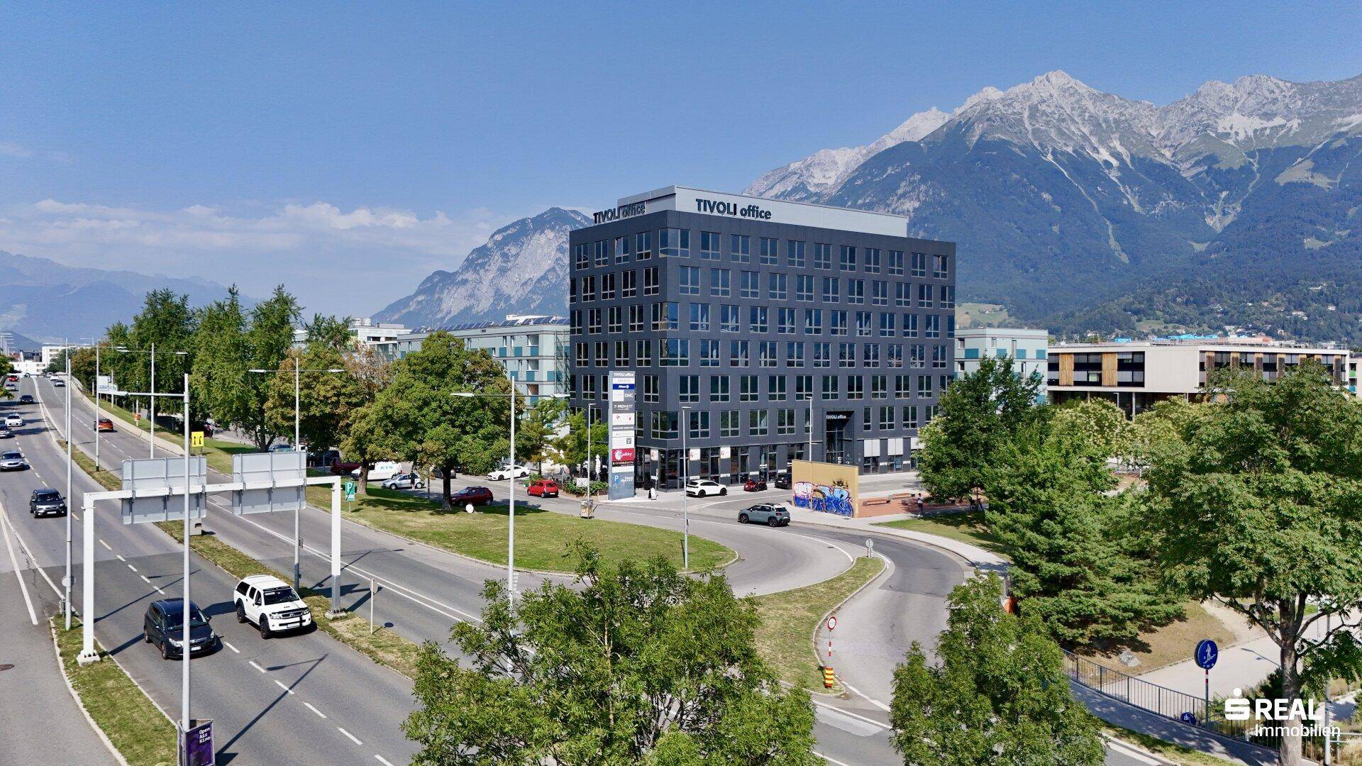 Luftaufnahme des modernen Bürogebäudes an einer Hauptstraße mit umliegender Bebauung und Bergpanorama.