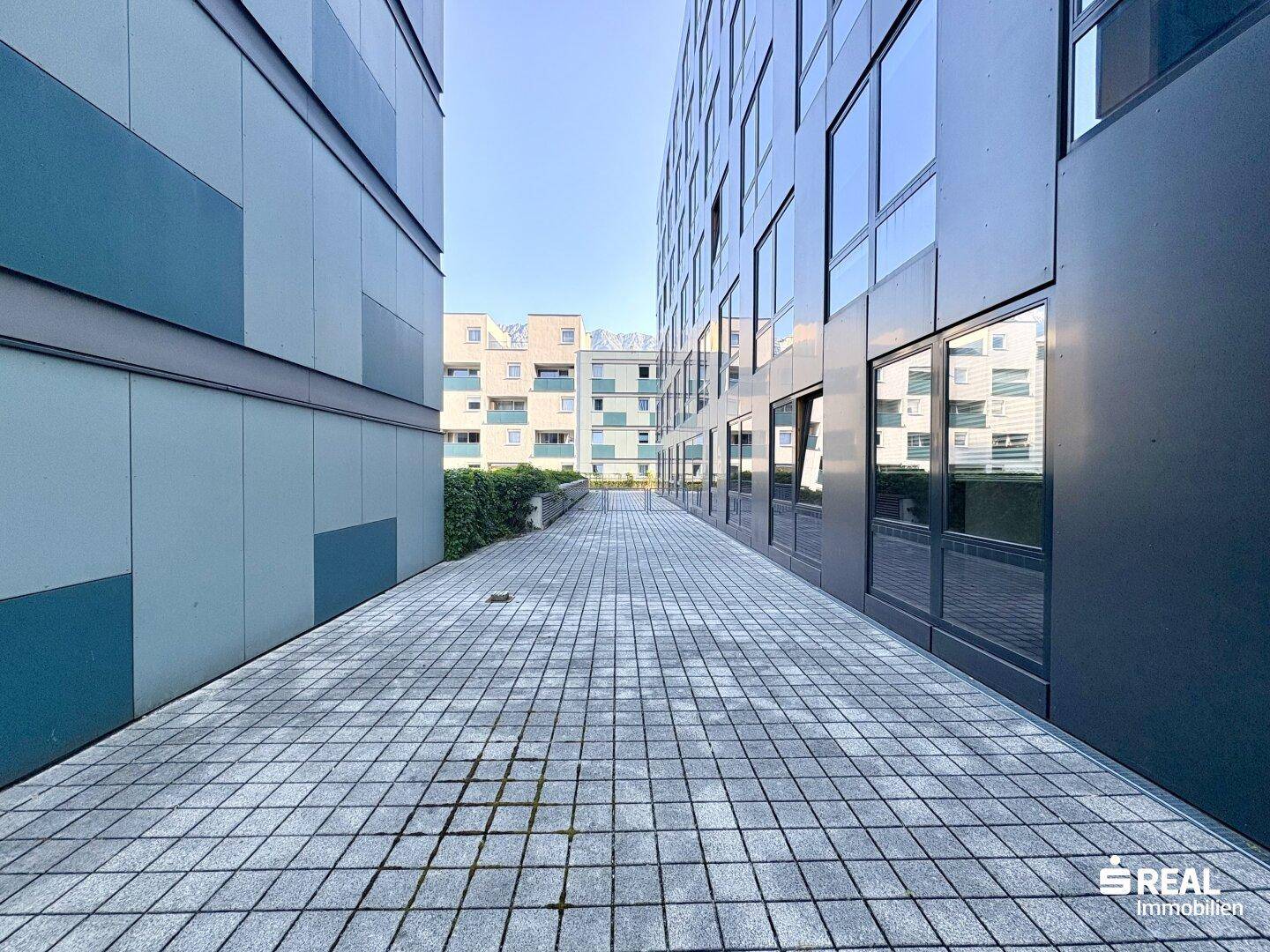 Gepflasterter Außenbereich zwischen zwei modernen Gebäuden mit großen Fenstern und schlichter Fassade.