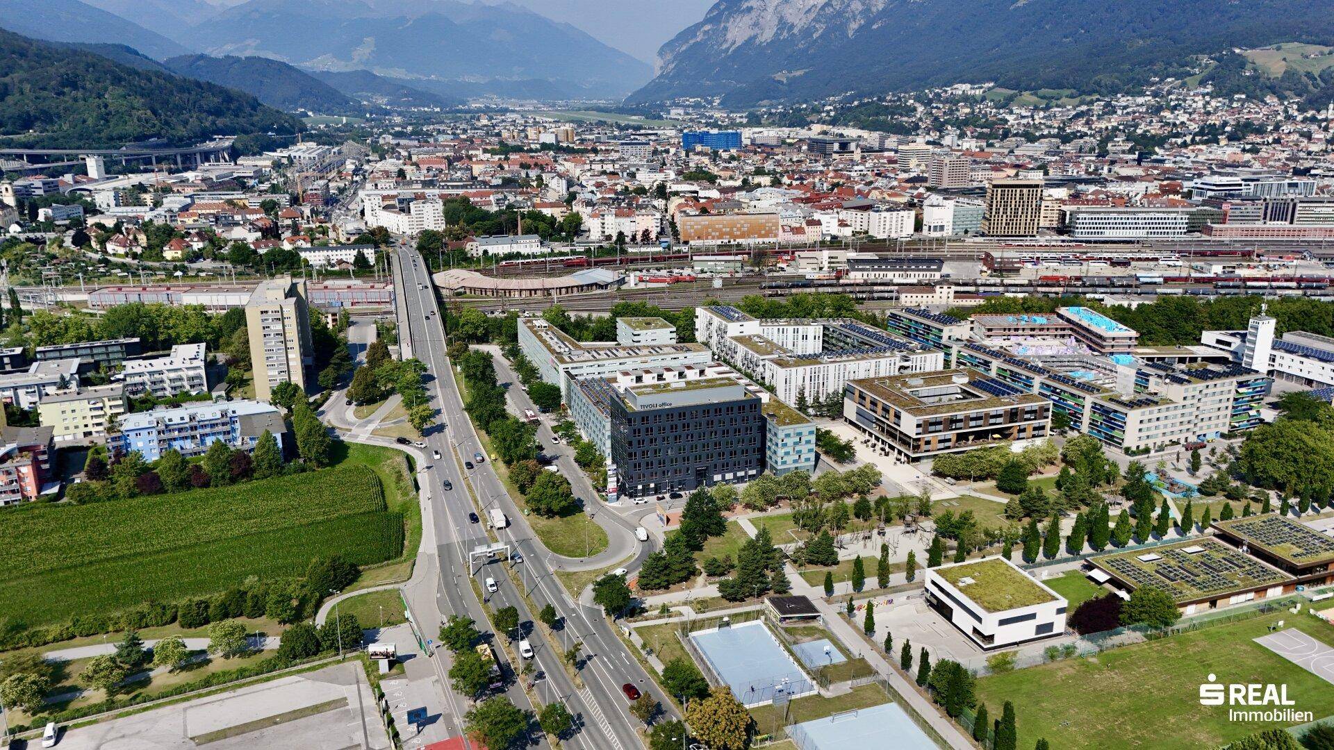 Weitläufige Luftaufnahme der Stadtlandschaft mit dem Bürokomplex, umgeben von Grünflächen und Bergen.