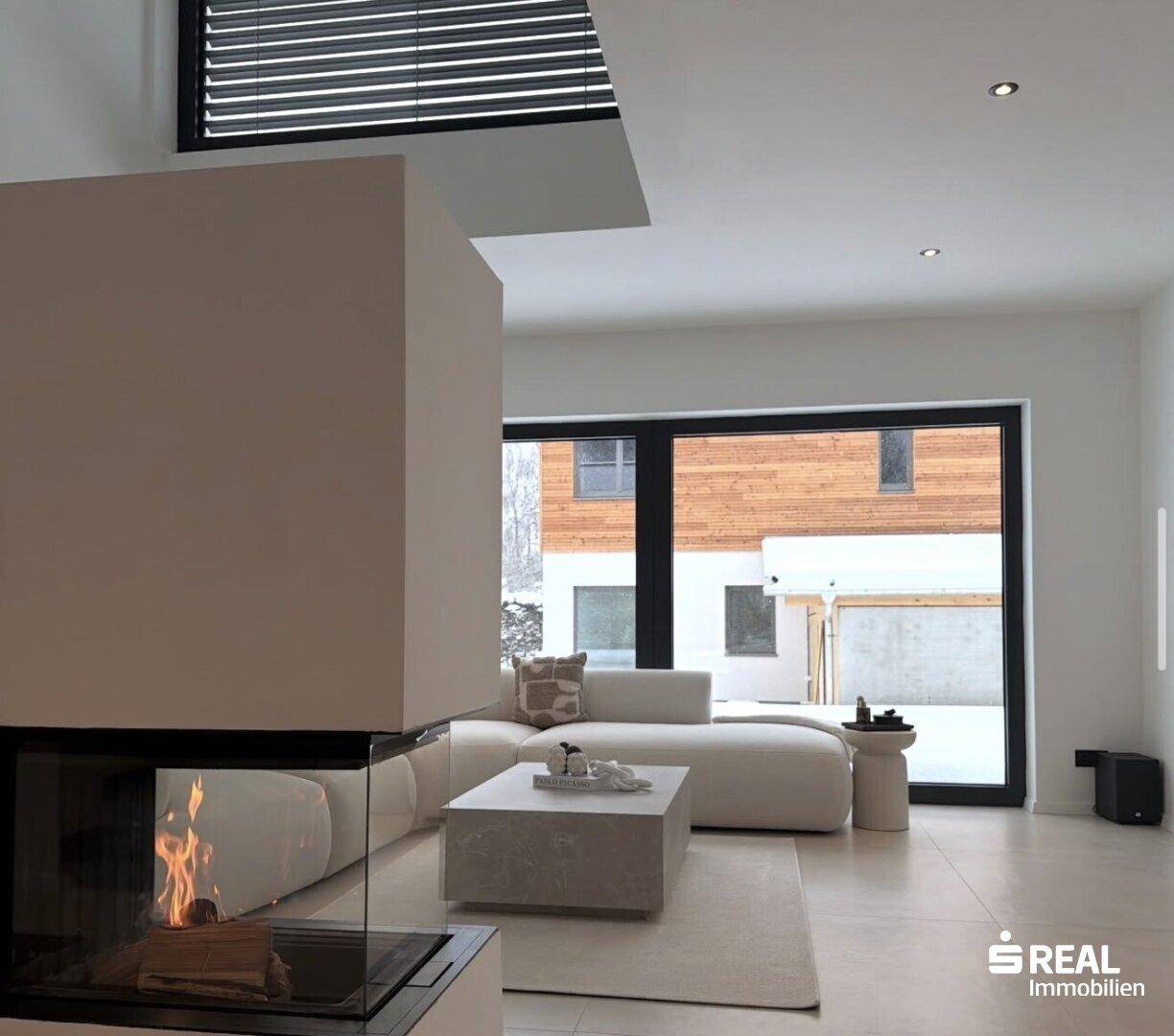 Gemütliches Wohnzimmer mit brennendem Kamin, Ecksofa und Blick ins Grüne durch große Fenster.