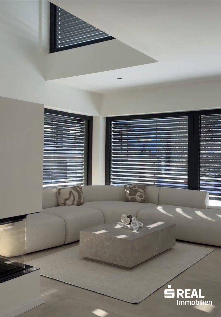 Helles Wohnzimmer mit großem Ecksofa, modernem Couchtisch und großen Fenstern mit Jalousien.