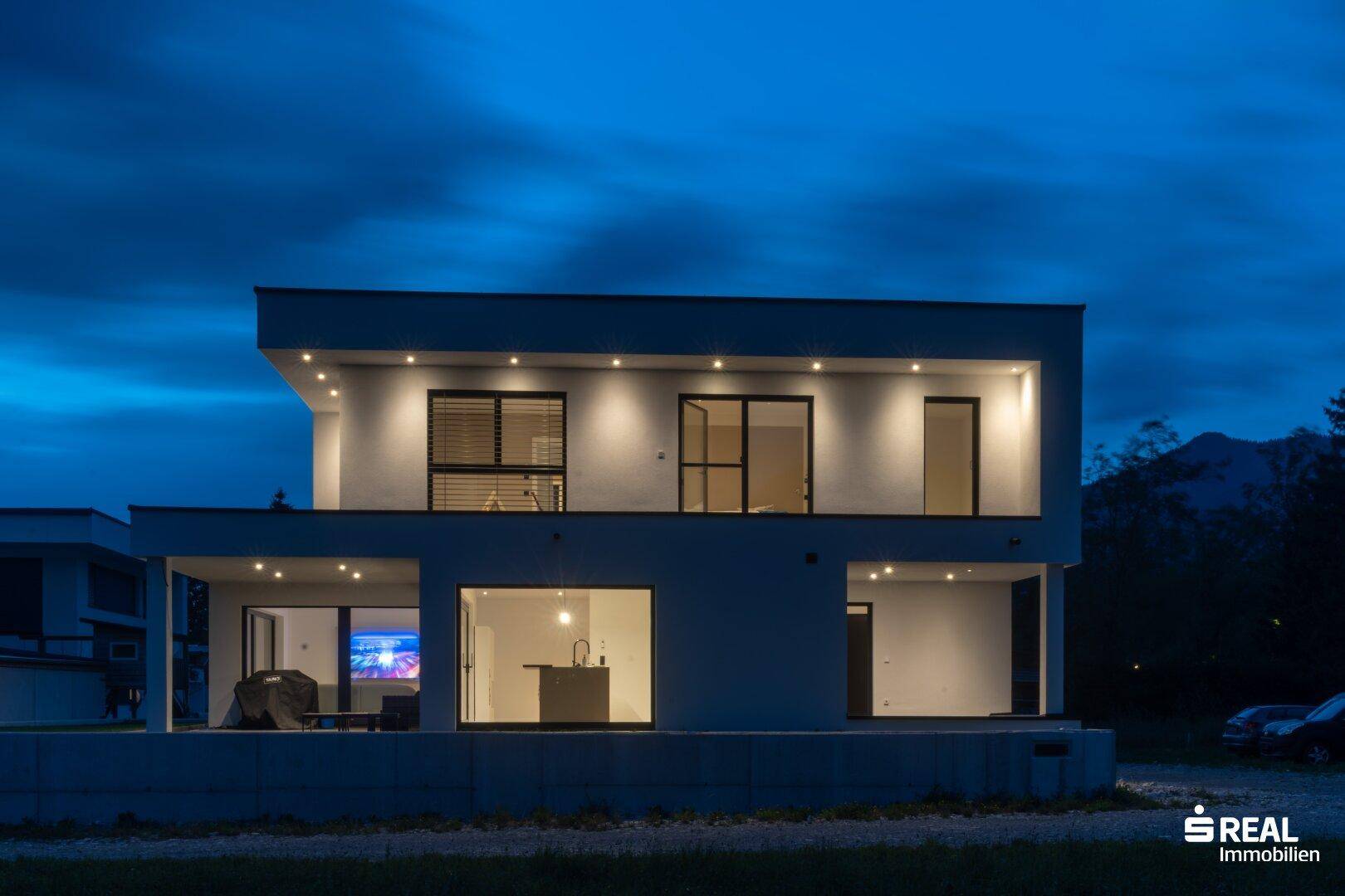Modernes Haus bei Nacht mit beleuchteten Innenräumen und großen Fensterfronten.