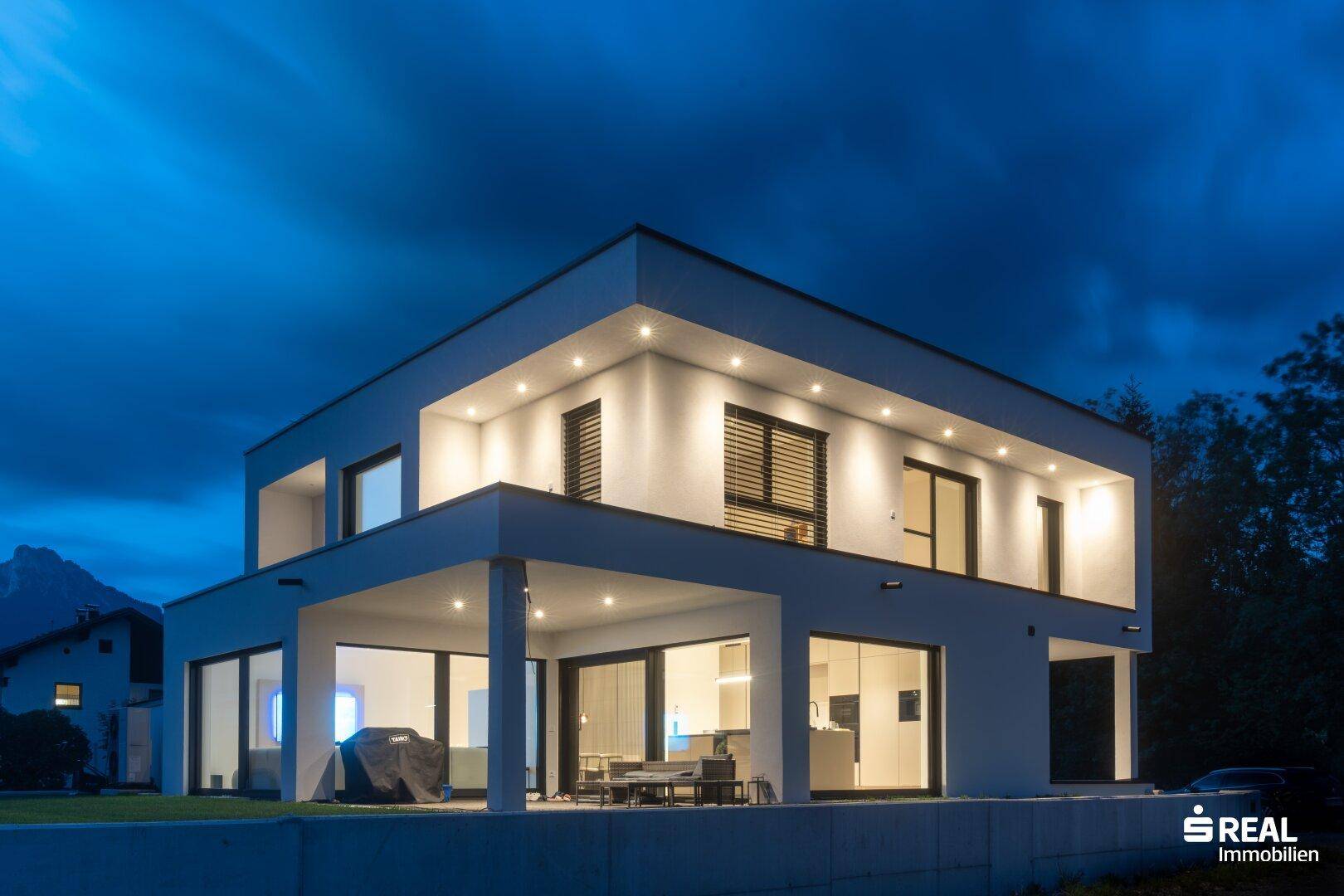 Modernes Haus bei Nacht mit beleuchteter Terrasse und großen Fensterfronten.
