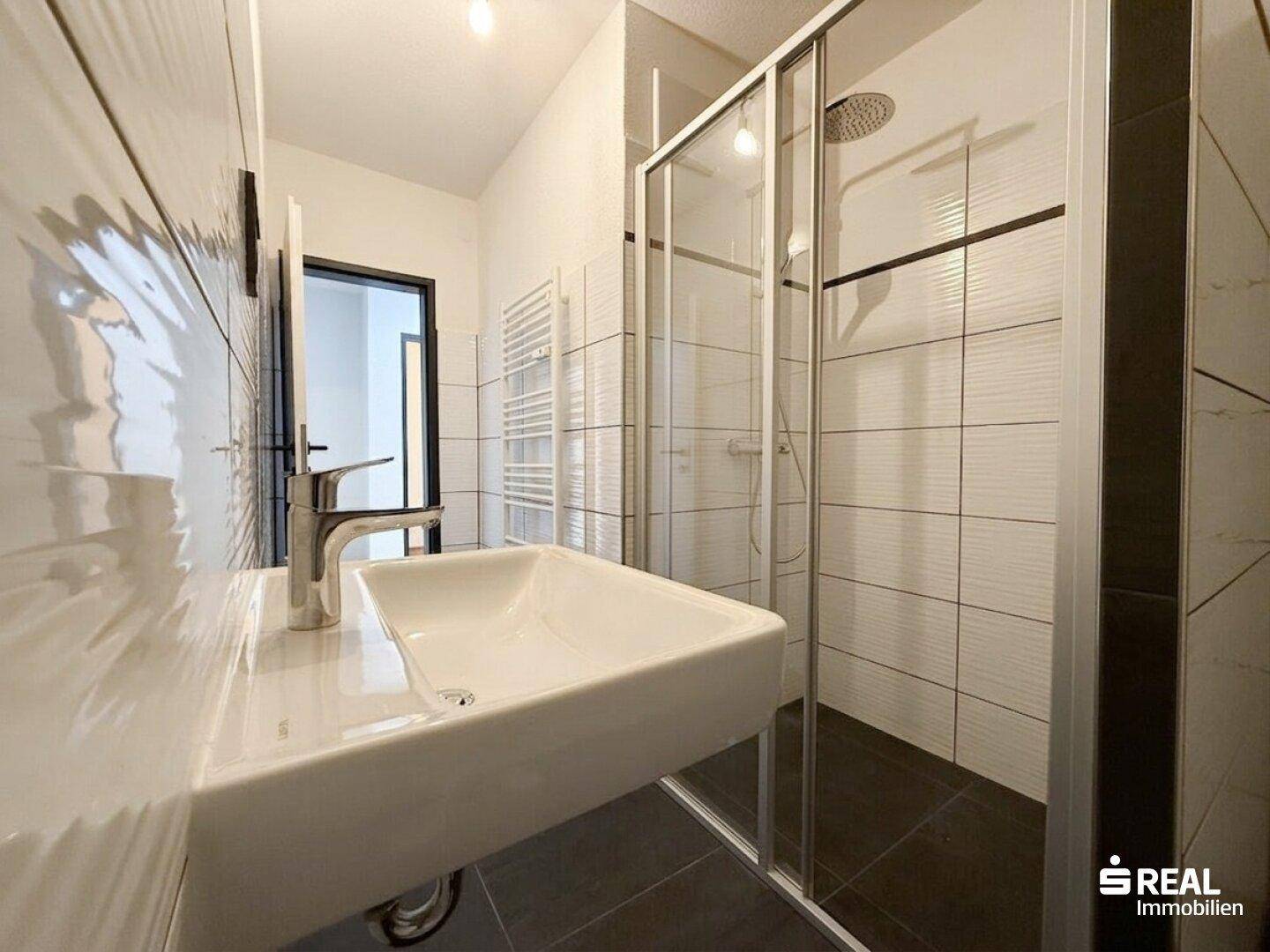 Modernes Badezimmer mit weißem Waschbecken, Duschkabine und dunklen Bodenfliesen.