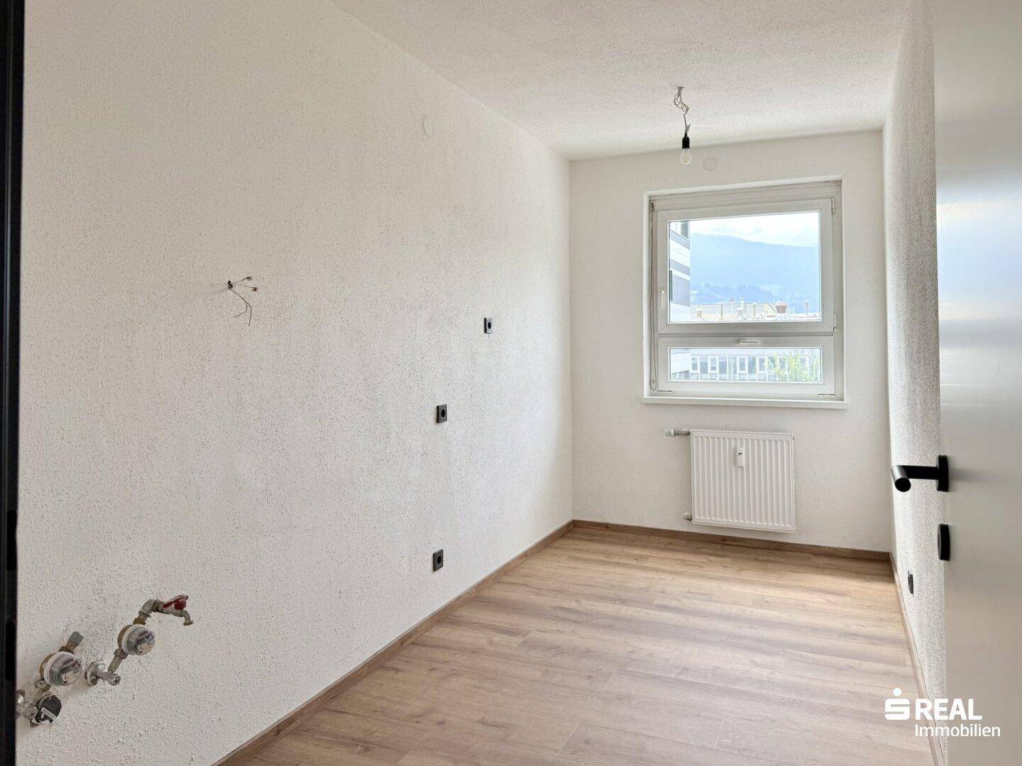 Leerer Raum mit sichtbaren Sanitäranschlüssen, Laminatboden und Fenster mit Bergblick.