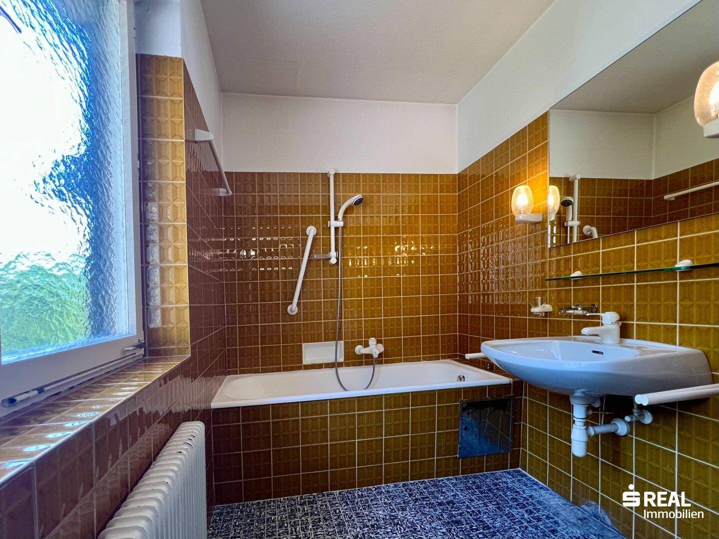 Badezimmer mit braunen Fliesen, Badewanne, Waschbecken und Fenster.