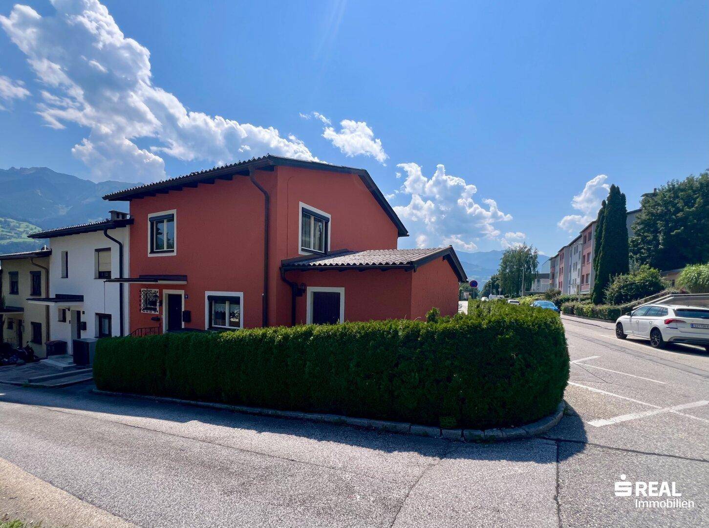 Rote Hausfassade mit Hecke und Straße, umgeben von Bergen und blauem Himmel.