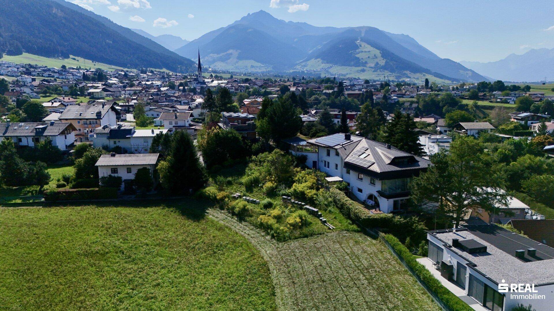 Malerische Luftaufnahme einer Wohngegend mit grünen Feldern und beeindruckendem Bergpanorama im Hintergrund.