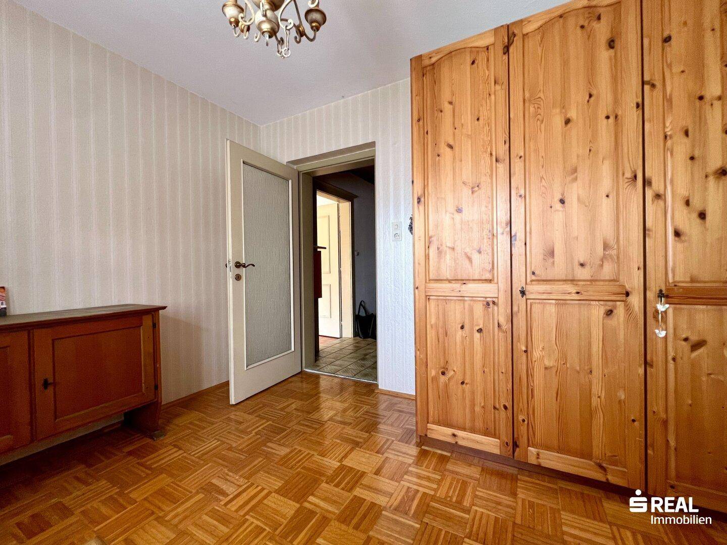 Schlafzimmer mit Holzboden, Holzschrank und Zugang zu einem weiteren Raum.