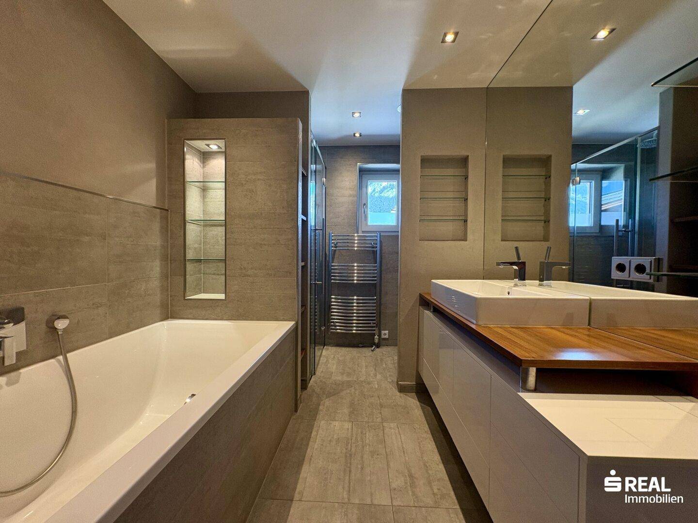 Modernes Badezimmer mit Badewanne, Doppelwaschbecken, Holzarbeitsplatte und begehbarer Dusche, hell gefliest.