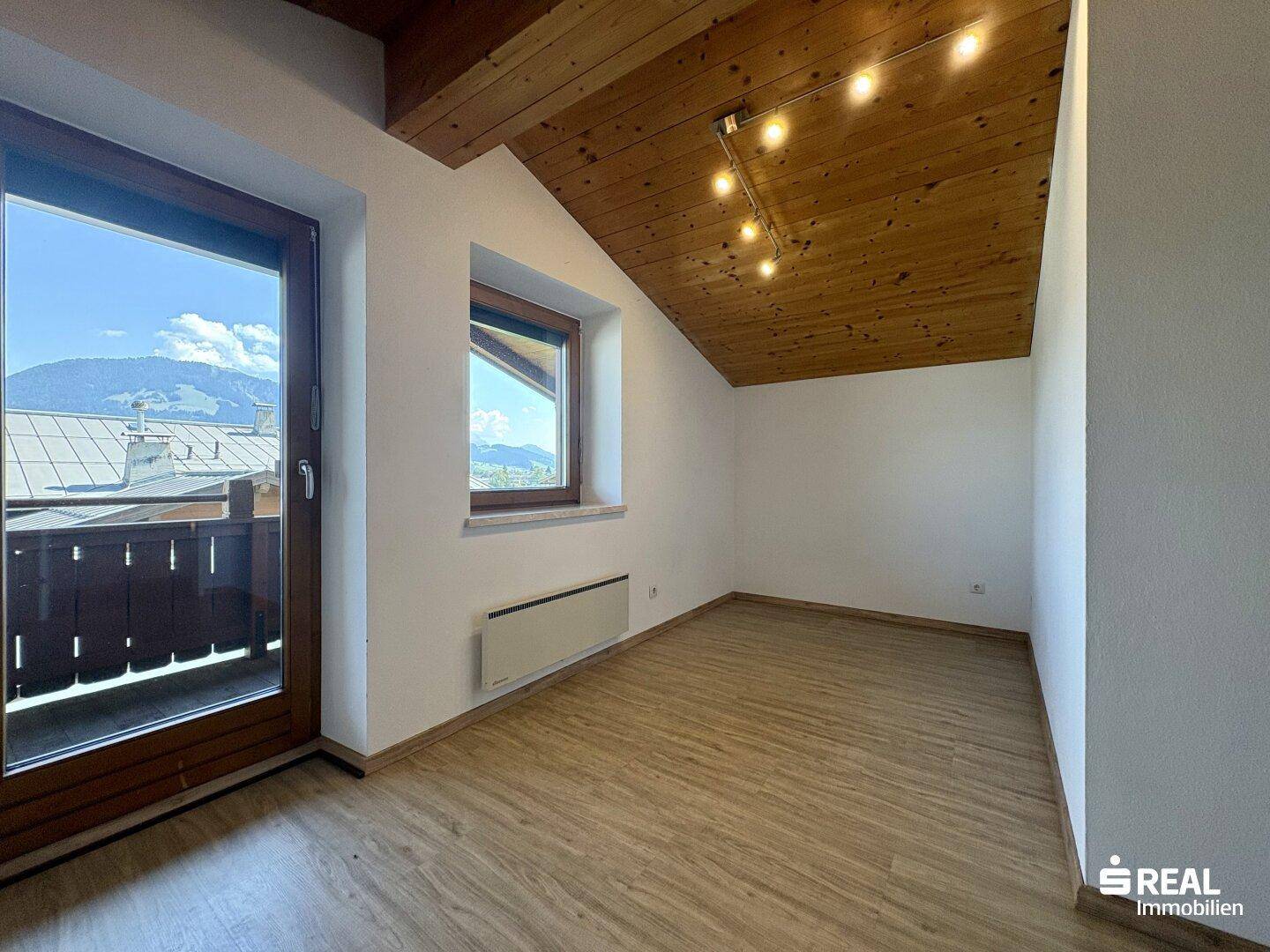 Helles Zimmer mit schräger Holzdecke, Laminatboden und Balkonzugang mit Bergblick.