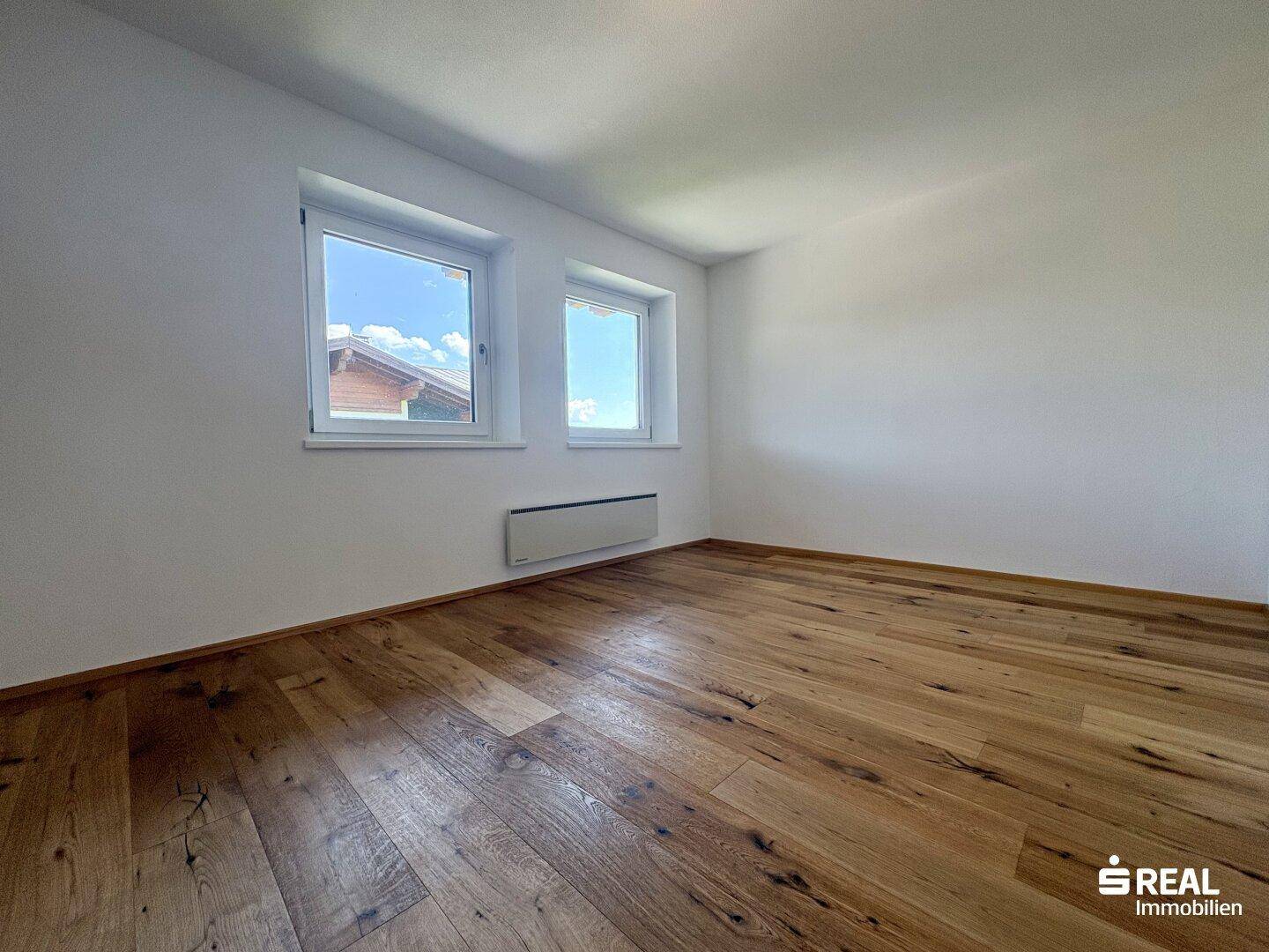 Helles Zimmer mit weiß gestrichenen Wänden, Holzboden und zwei Fenstern mit Blick nach draußen.