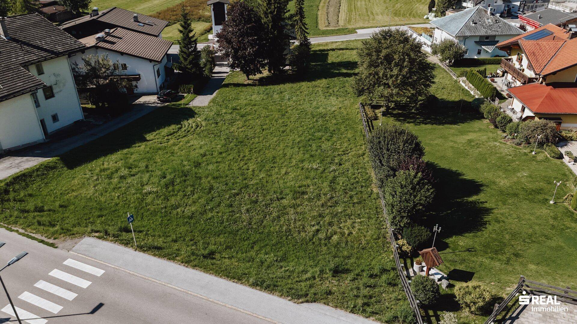 Grünes Baugrundstück an einer Straße mit Hecke und Blick auf die umliegenden Häuser.