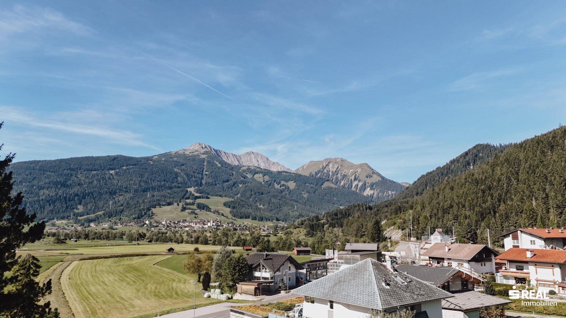 Malerischer Blick auf ein Dorf, grüne Felder und bewaldete Berge unter blauem Himmel.