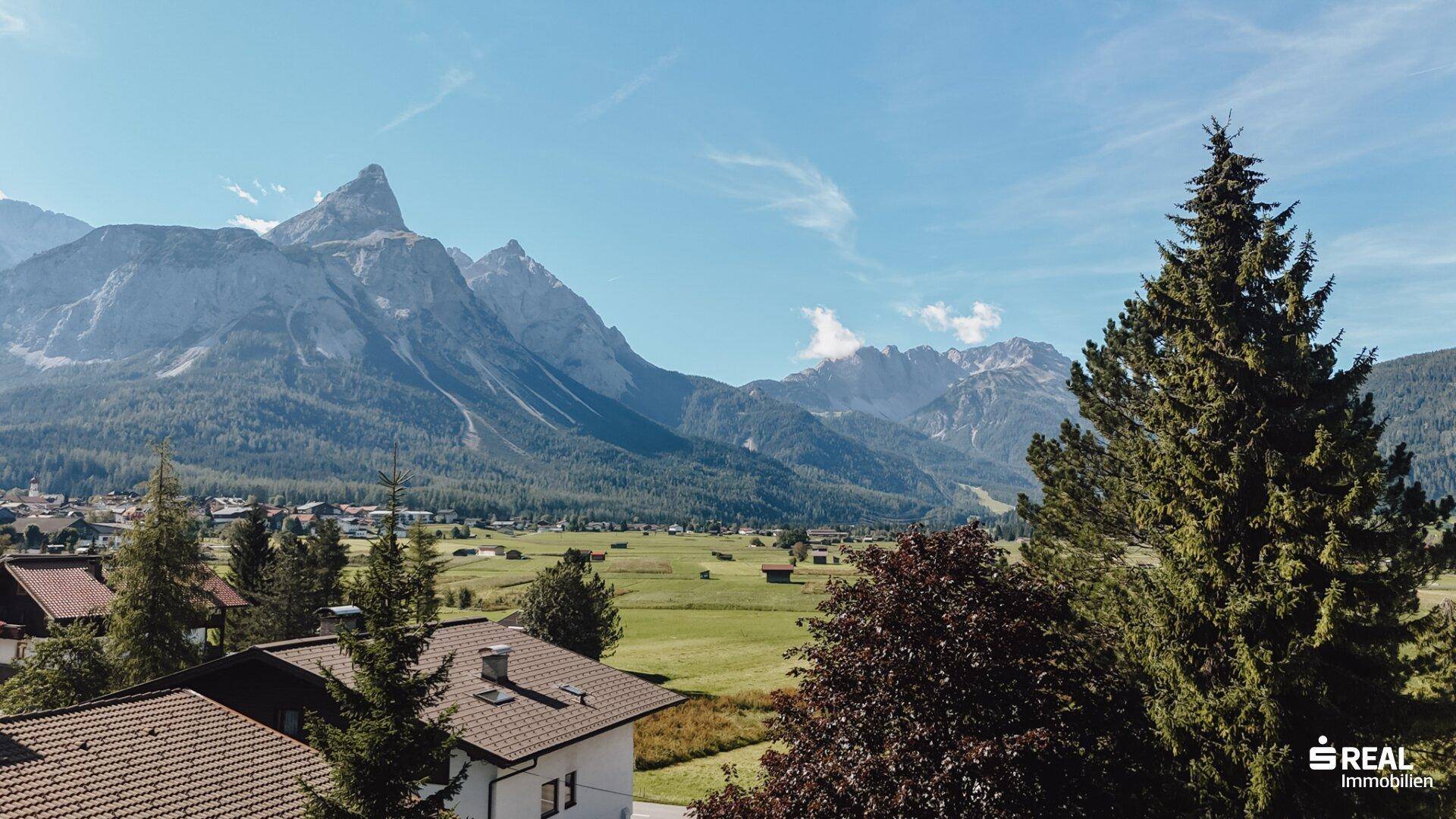 Weitblick über ein Dorf mit grünen Feldern und beeindruckenden Berggipfeln unter klarem Himmel.