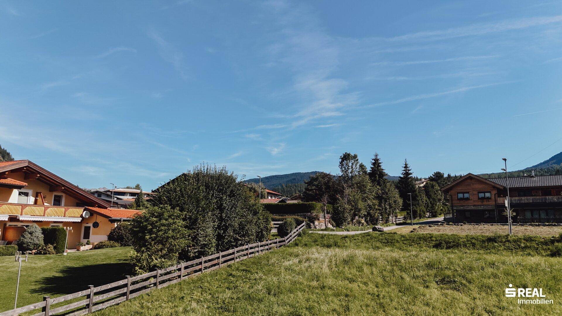 Grünes Grundstück mit Holzzaun und Blick auf umliegende Häuser und bewaldete Hügel.