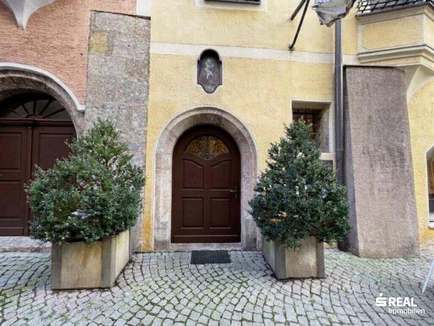 Historische Fassade mit Holztür und gepflastertem Eingangsbereich, umgeben von alten Gebäuden.