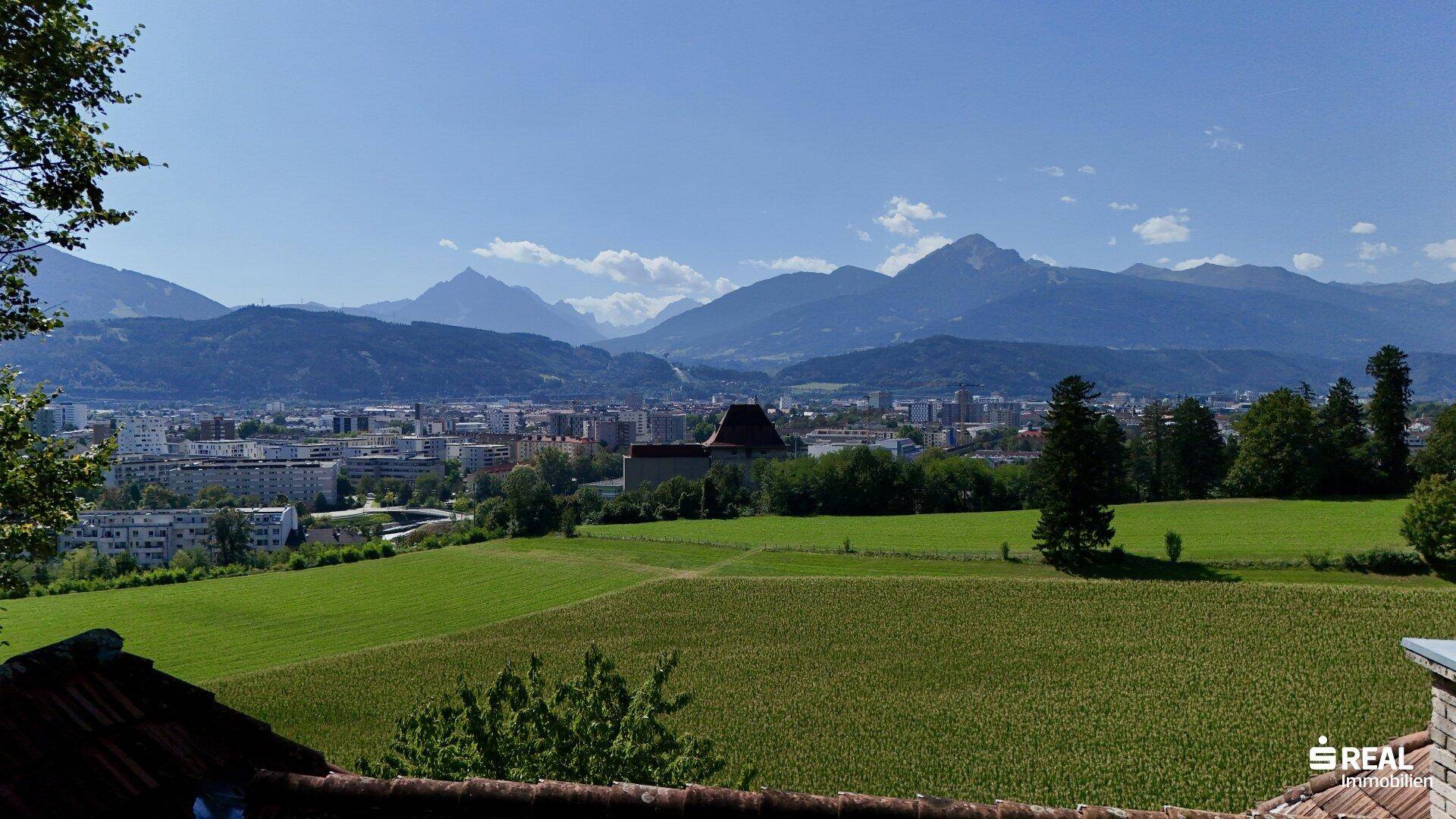 Weitläufiger Blick über grüne Felder, eine Stadt und majestätische Berge im Hintergrund bei Tag.