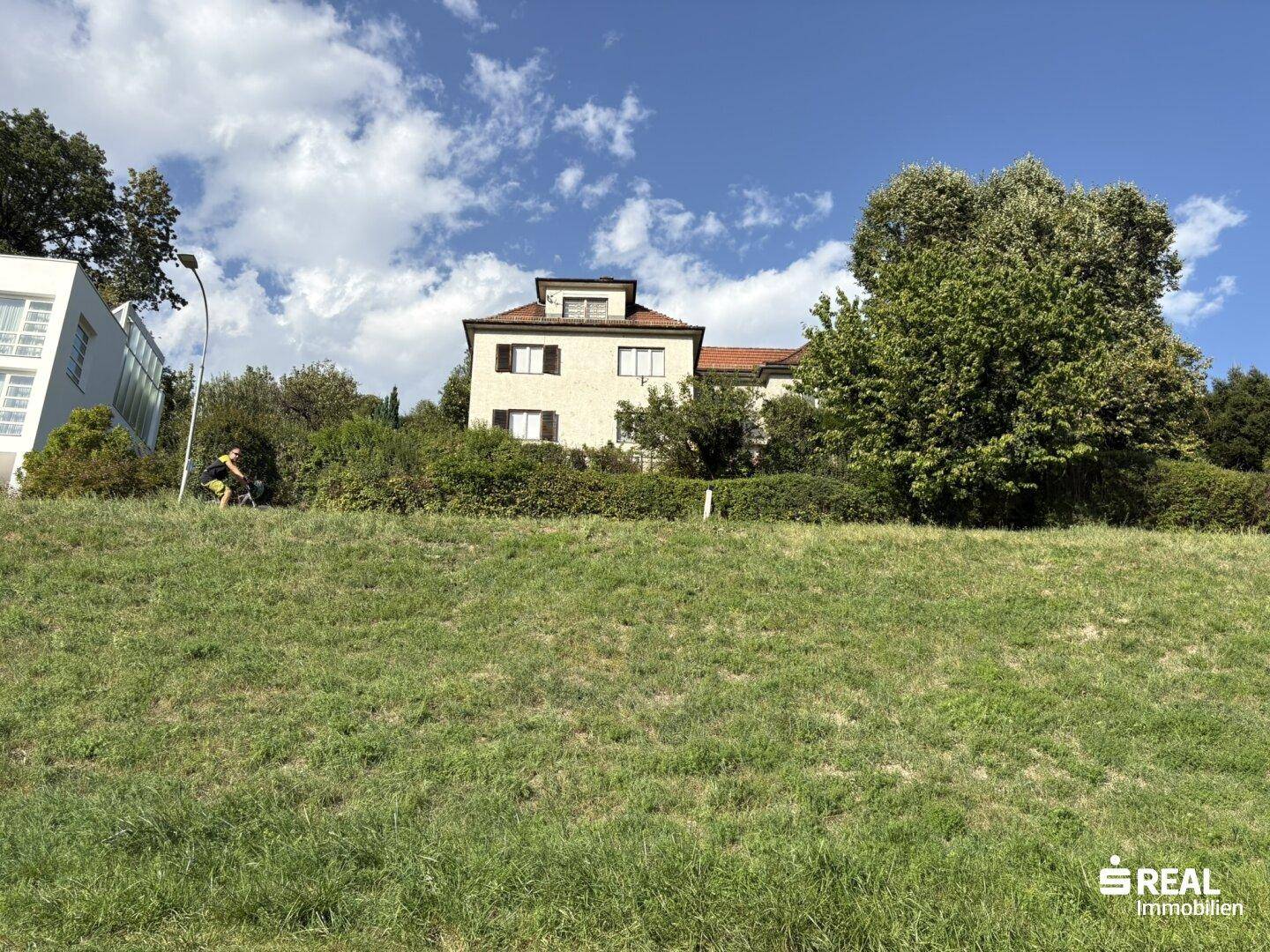Ein traditionelles Haus auf einem grünen Hügel, umgeben von Bäumen und Büschen unter blauem Himmel.