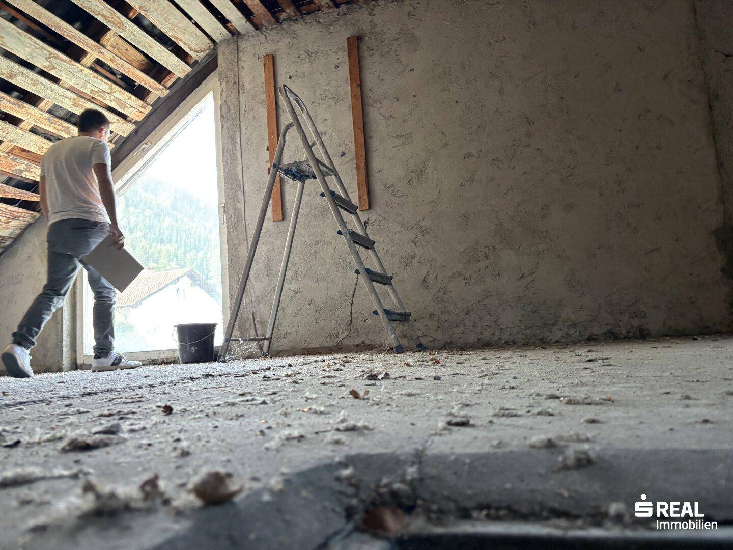 Rohbau-Dachboden mit einem Arbeiter, einer Leiter und einem großen Dachfenster, das viel Tageslicht hereinlässt.
