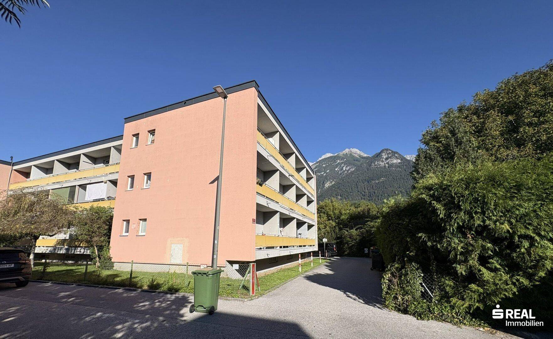 Ansicht des Mehrfamilienhauses mit Balkonen, umgeben von Bäumen und Bergen im Hintergrund.