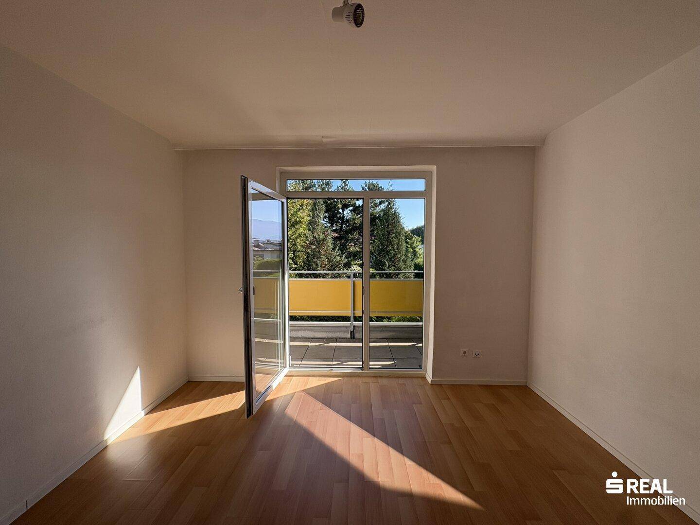 Helles Zimmer mit Holzboden und Zugang zu einem Balkon, durchflutet von Sonnenlicht.