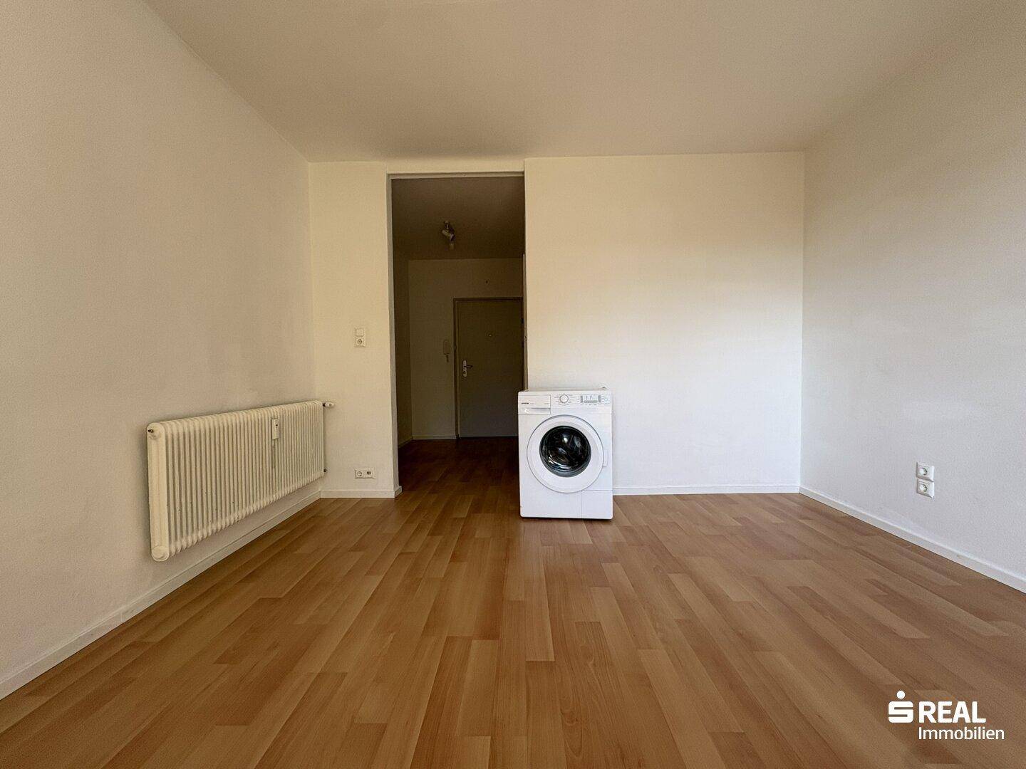 Heller Raum mit Holzboden, Heizkörper und Waschmaschine, führt zu einem weiteren Bereich.