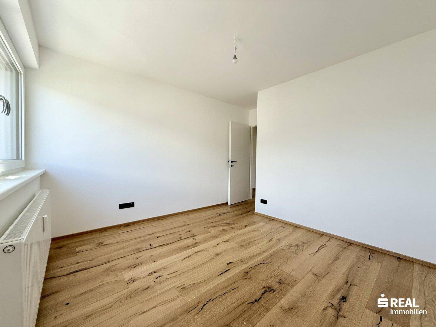 Leeres Zimmer mit Holzboden und einem Fenster, das viel Tageslicht hereinlässt.