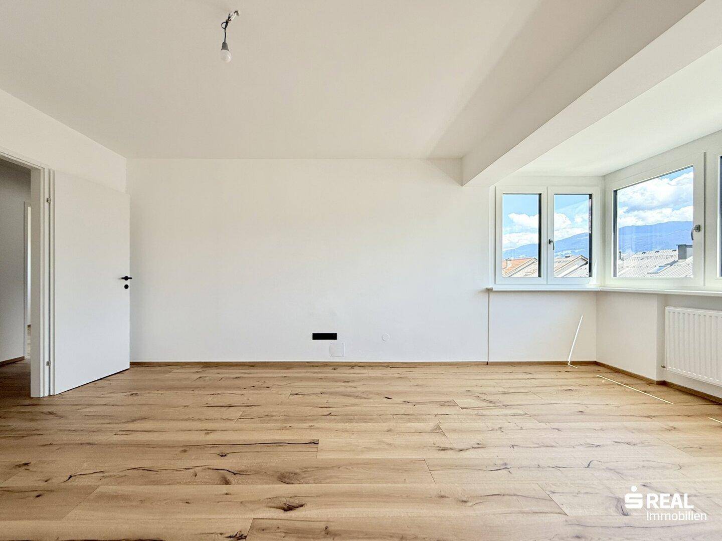 Heller, leerer Raum mit Holzboden und einer Fensterfront, die einen weiten Ausblick ermöglicht.