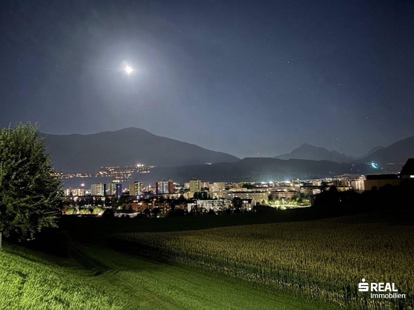 Nachtansicht einer beleuchteten Stadt vor einer Bergkulisse unter einem klaren Sternenhimmel.