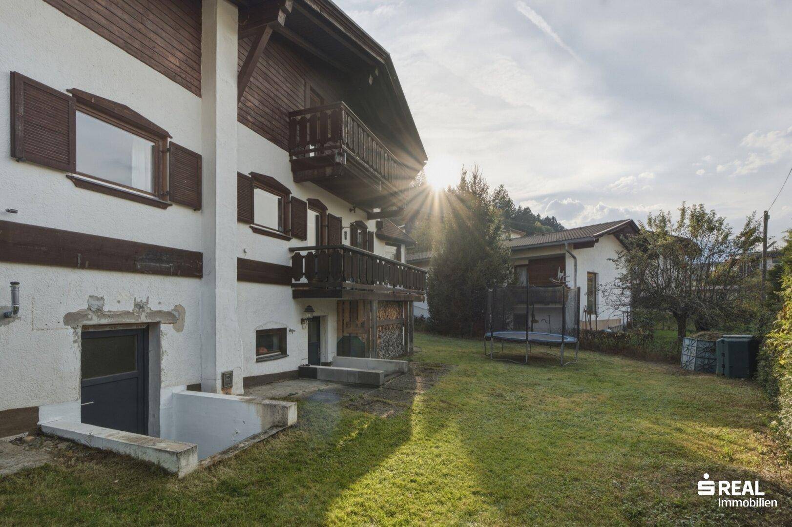 Hausfassade mit Holzbalkonen und einem sonnigen Gartenbereich mit Trampolin.