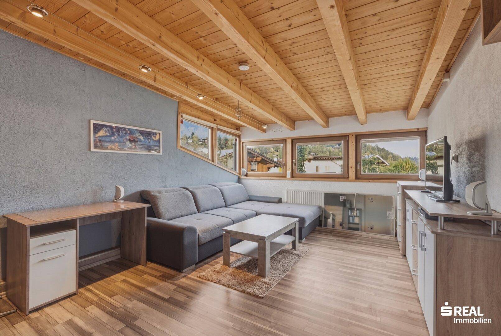 Gemütliches Wohnzimmer mit Holzbalkendecke, Sofa und Blick auf die grüne Umgebung.