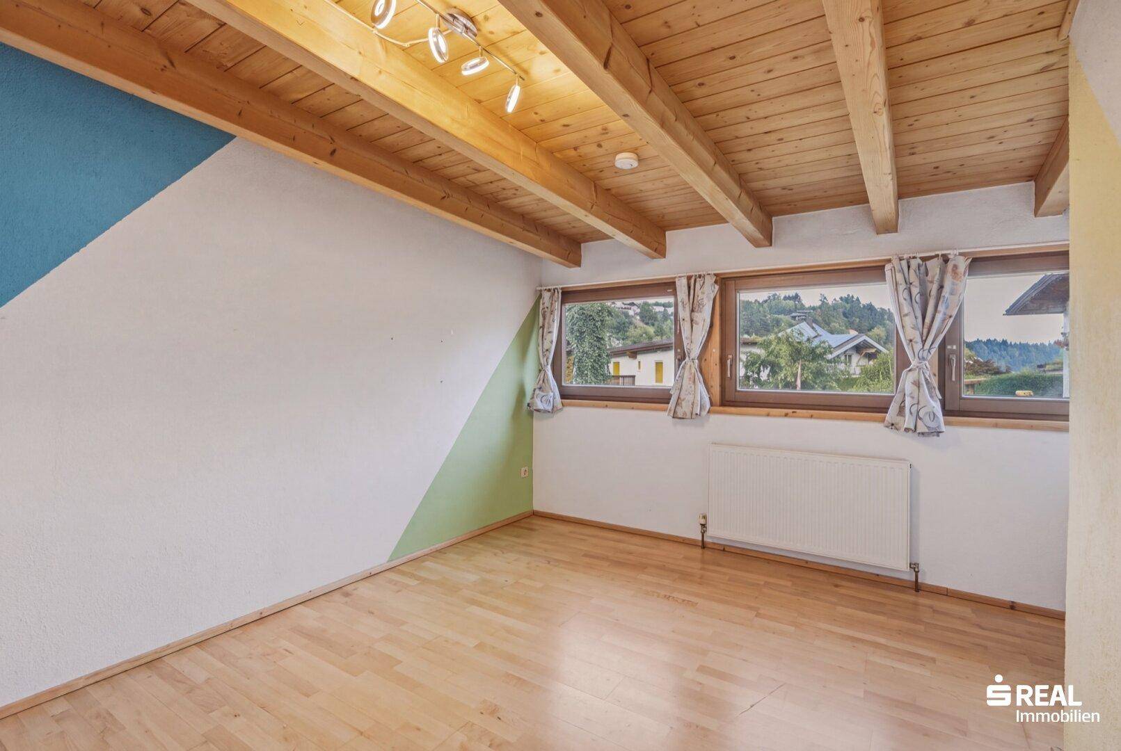 Helles Zimmer mit Holzbalkendecke, Parkettboden und Fenstern mit Blick ins Grüne.