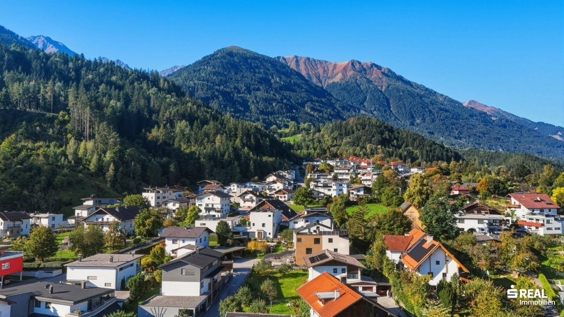 Luftaufnahme einer Wohnsiedlung am Fuße eines bewaldeten Berges unter blauem Himmel.
