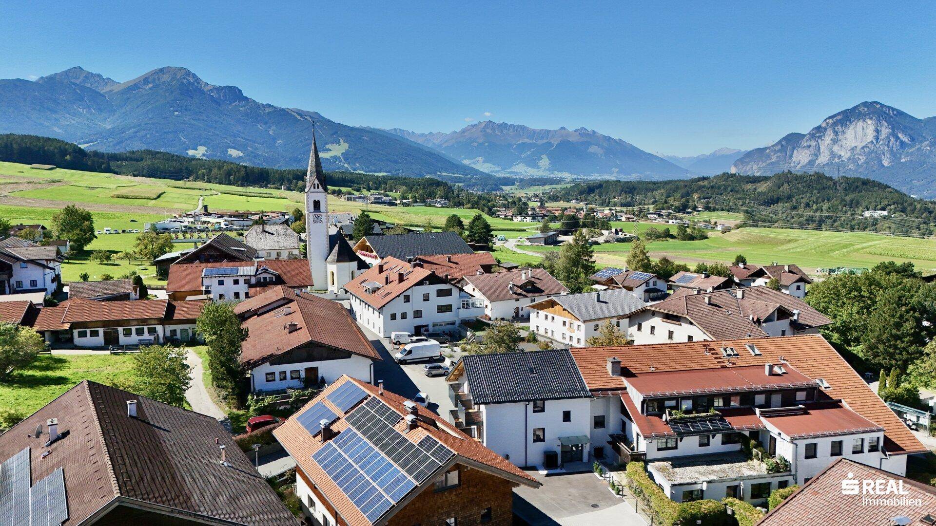 Panoramablick auf ein idyllisches Dorf mit traditionellen Häusern und einer Kirche vor alpiner Kulisse.