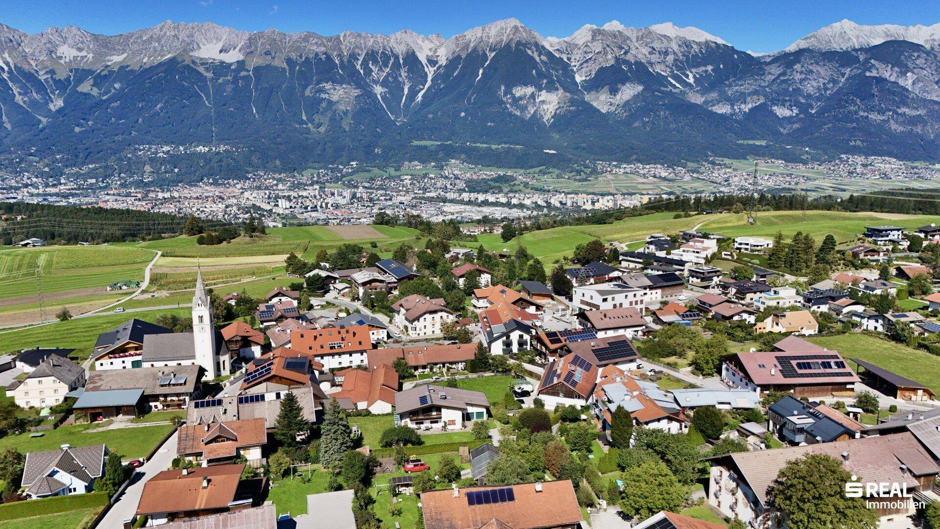 Weitwinkelaufnahme eines charmanten Dorfes, eingebettet in eine grüne Landschaft mit hohen Bergen im Hintergrund.