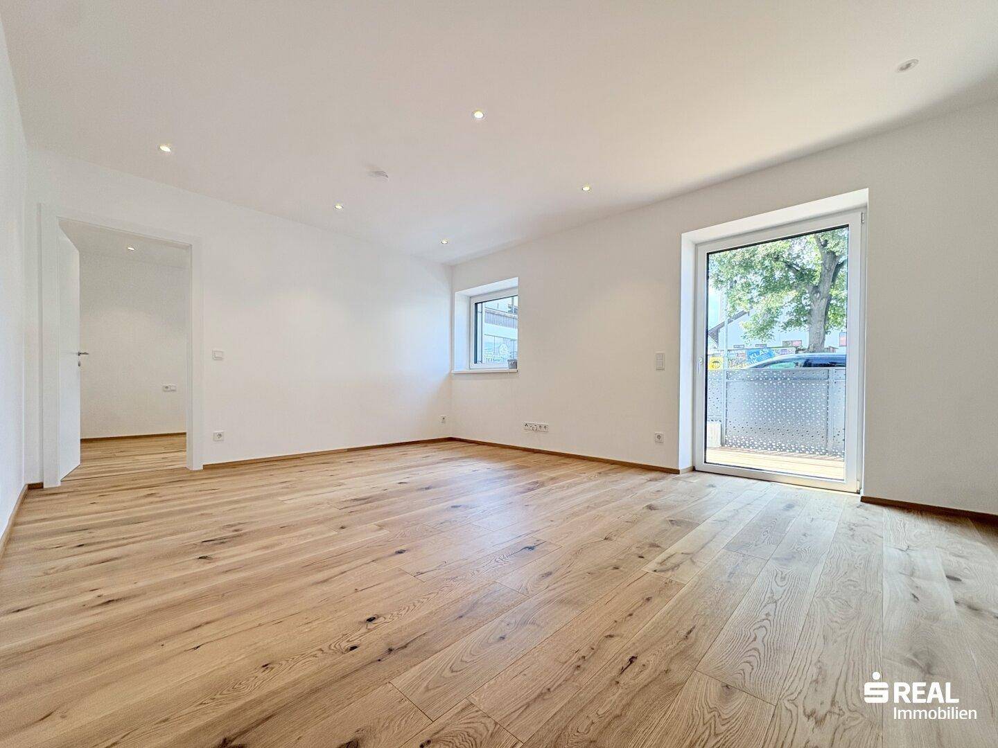 Heller, leerer Wohnraum mit neuem Holzboden, weißen Wänden und Zugang zu einem Balkon.