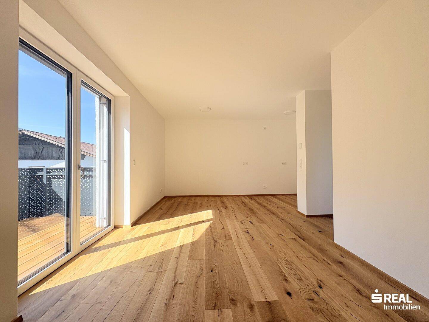 Geräumiger, unmöblierter Wohnbereich mit hellem Holzboden und großen Schiebetüren zum Balkon.