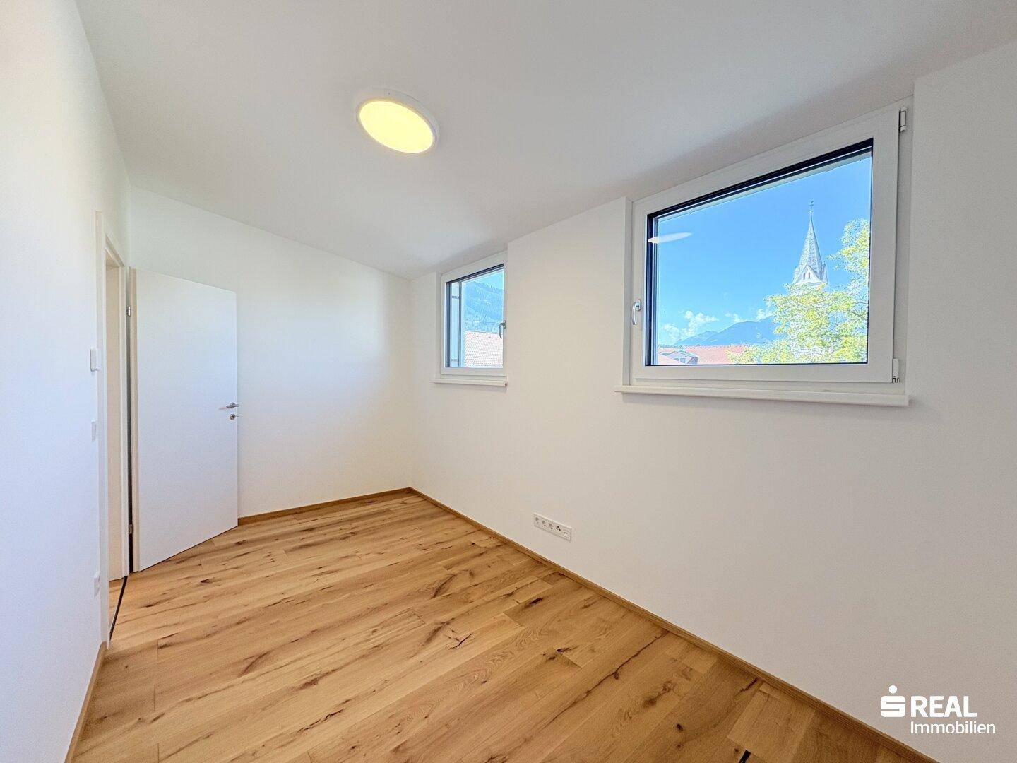 Helles Schlafzimmer mit Holzboden, weißen Wänden und Fenstern mit Blick auf eine Kirche und Berge.