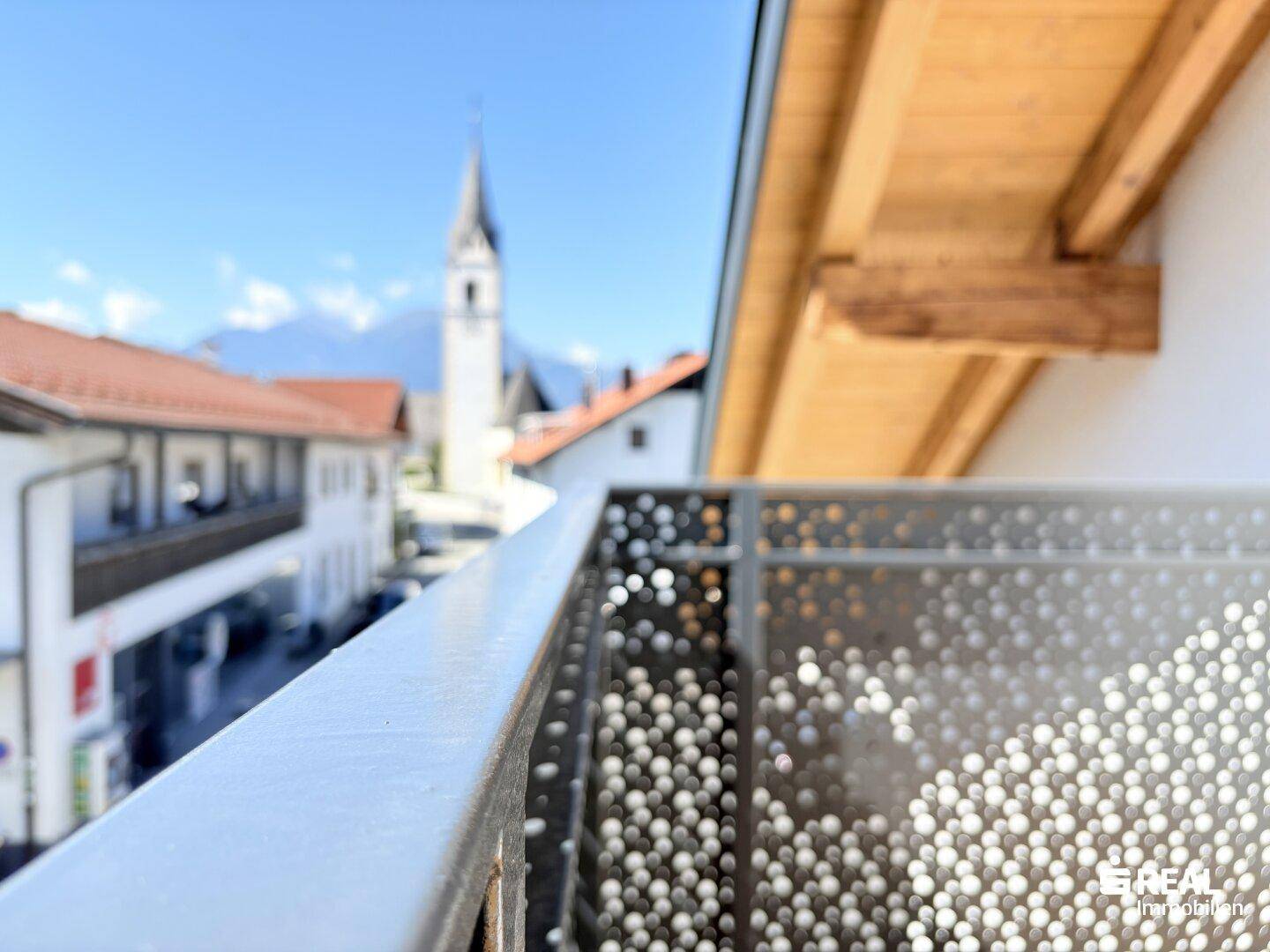 Nahaufnahme eines Balkongeländers mit verschwommener Kirchturmspitze und Bergen im Hintergrund.