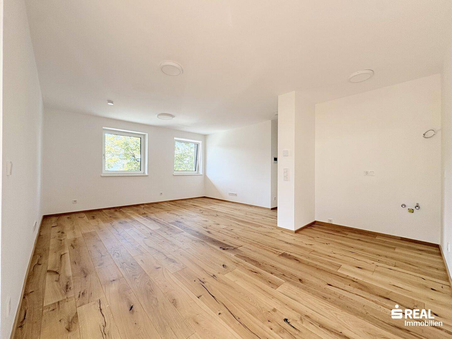 Großer, heller Raum mit Holzboden und zwei Fenstern, ideal für einen offenen Wohnbereich.