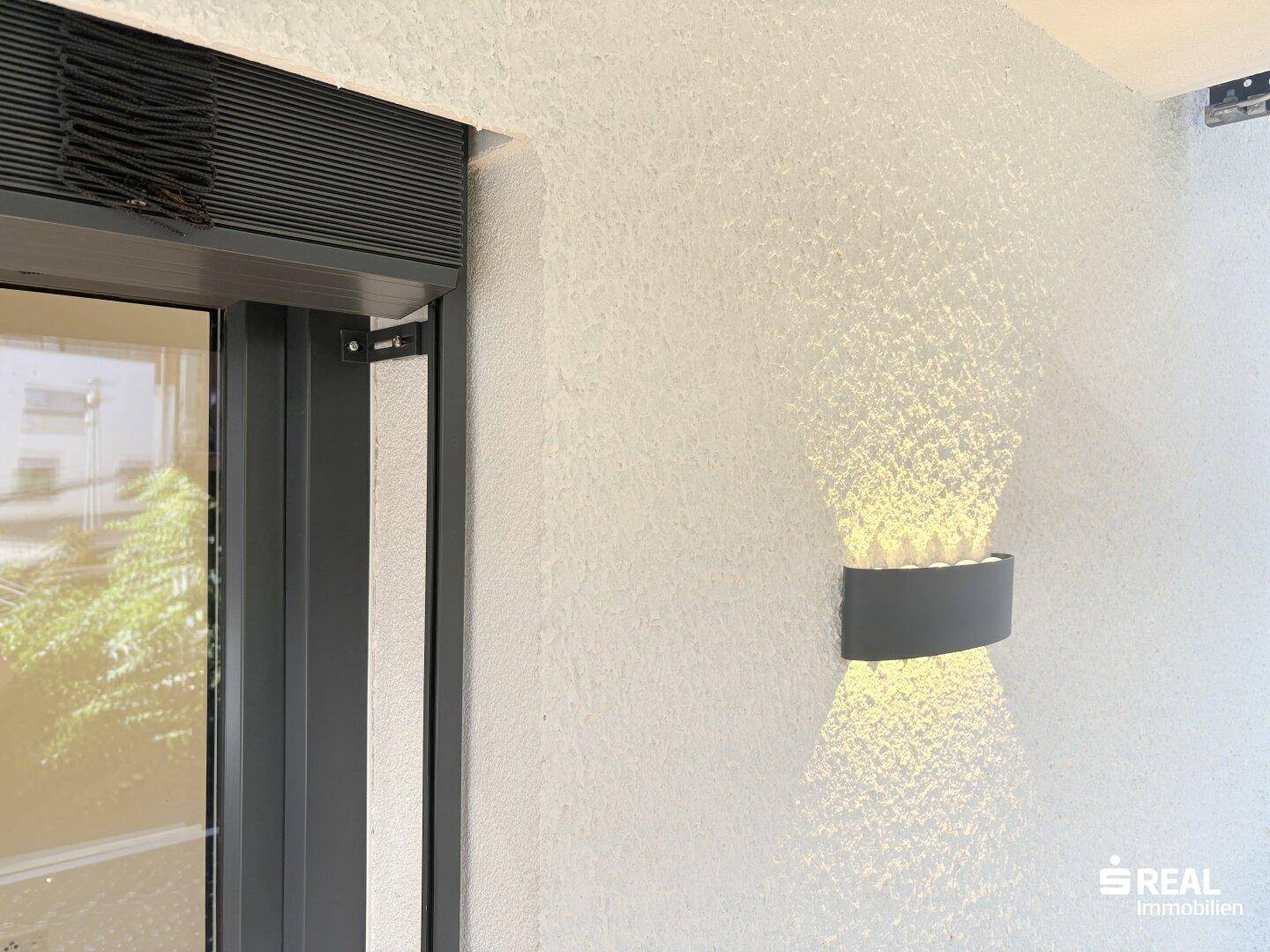 Außenwand mit moderner Beleuchtung und einem Fenster mit integriertem Rollladen für Privatsphäre.