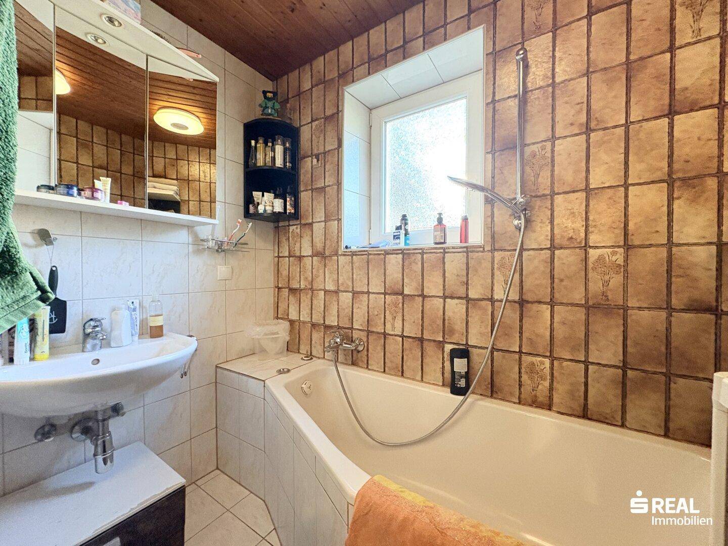 Badezimmer mit Badewanne, Waschbecken, Spiegelschrank und Fenster, gefliest in Brauntönen.