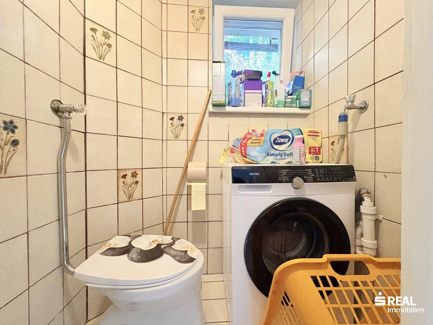 Kleines WC mit Waschmaschine und Fenster, gefliest und funktional eingerichtet.