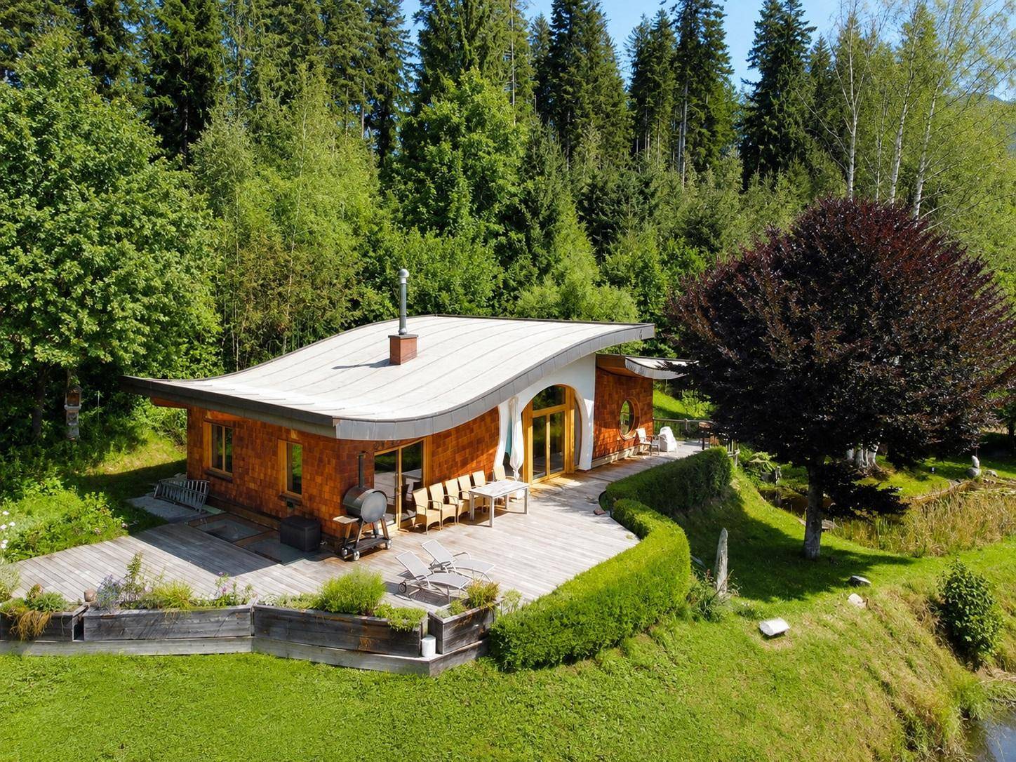 Vogelperspektive auf ein modernes Holzhaus mit geschwungenem Dach und großer Holzterrasse inmitten grüner Natur.