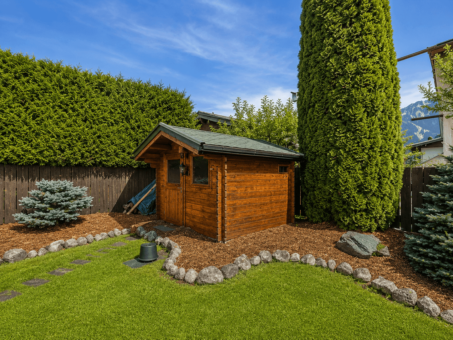 Rustikales Gartenhaus aus Holz, umgeben von gepflegtem Rasen und hohen Hecken.